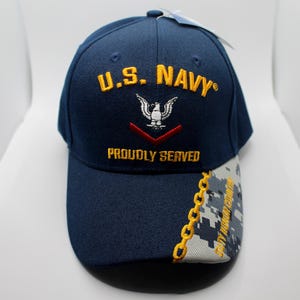 Könnte beinhalten: Marineblaue Baseballkappe mit gesticktem Text und Grafiken. Auf der Vorderseite steht "U.S. NAVY" in Gold, ein weißes Adleremblem und "PROUDLY SERVED". Der Schirm hat ein Tarnmuster.