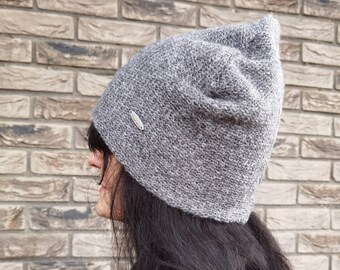 May include: A gray knit beanie with a small silver tag. The hat is worn by a person with long, dark hair. The beanie has a textured appearance and a slightly pointed top. The background is a brick wall.