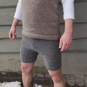 May include: A knitted brown vest and grey shorts. The vest is sleeveless with a ribbed hem. The shorts are knee-length and form-fitting. The person is wearing a white long-sleeved shirt under the vest. The clothing appears to be made of wool.