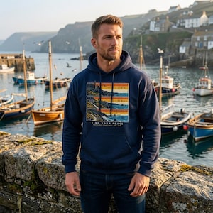 Puede incluir: Una sudadera con capucha azul marino que presenta un estampado gráfico de tres paneles de paisajes costeros apilados con el texto CORN WALL OVERNOW y FIND YOUR PEACE SOUTH WEST COAST PATH - EST. 1978 en el pecho.