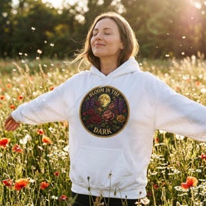 Puede incluir: Sudadera con capucha blanca con bolsillo delantero y un diseño gráfico circular. El diseño presenta una luna llena, rosas y el texto "BLOOM IN THE DARK". La sudadera con capucha se usa en un campo de flores silvestres.