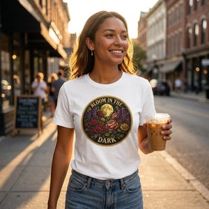 Puede incluir: Camiseta blanca con un gráfico circular que dice "Bloom in the Dark". El gráfico presenta una luna llena, rosas y otras flores. La persona lleva vaqueros azules y sostiene una bebida.