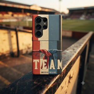 Puede incluir: Una funda para smartphone con un diseño tricolor vertical en rojo, crema y azul oscuro. El centro muestra dos bufandas anudadas y el texto "TEAM" en letras blancas grandes, con "UNITED BY THE GAME" impreso en letra más pequeña debajo.