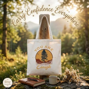 Puede incluir: Bolso tote color crema con el texto "You Are Enough" y un gráfico de un árbol y montañas. Las palabras "Carry Confidence Everywhere" están arqueadas arriba. Un diario, un bolígrafo y una taza están en primer plano.