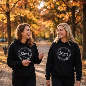 May include: Two people wearing black hoodies with the text "Bloom anyway" inside a wreath design. The hoodies have a drawstring hood and a front pocket. The background shows a park with trees and fall foliage.