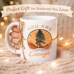 Puede incluir: Taza de cerámica blanca con las palabras "YOU ARE Enough" en letras doradas. La taza presenta un diseño circular con un árbol y un paisaje montañoso en tonos naranja y azul. Un lazo de cuerda está atado alrededor del asa.