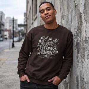 Puede incluir: Una sudadera marrón oscuro con el texto "Love Grows Here" en un diseño floral. El diseño incluye flores, un pájaro y raíces. La sudadera es de cuello redondo.