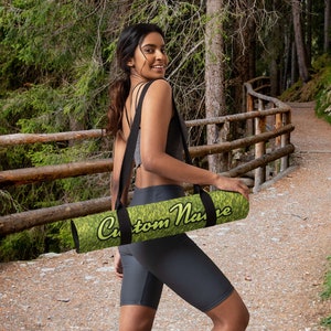 Puede incluir: Una mujer caminando por un sendero en un bosque llevando una esterilla de yoga verde y amarilla con una correa negra y el texto "Custom Name" impreso en ella.
