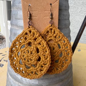 Puede incluir: Pendientes de ganchillo de color amarillo mostaza con forma de lágrima. Los pendientes se muestran en una tarjeta marrón con un gancho de metal. El diseño de ganchillo presenta un patrón abierto y circular. Los pendientes cuelgan de un recipiente de metal galvanizado.