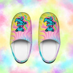 May include: A pair of colorful slippers with a tie-dye pattern in shades of yellow, pink, and blue. Each slipper features a teal bear graphic. The slippers have a white interior and a black trim.
