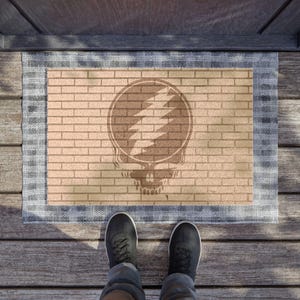 May include: A rectangular doormat with a brick-like pattern and a Steal Your Face skull logo. The mat has a light brown center with a dark brown design and a blue and white border. The image is taken from a top-down perspective.