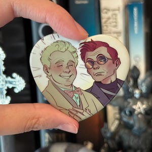 Könnte beinhalten: Herzförmiger Button mit zwei Zeichentrickfiguren. Einer hat blondes Haar, einen beigen Anzug und eine rosa Fliege. Der andere hat rotes Haar, eine Brille und einen lila Anzug. Der Button wird in einer Hand gehalten.