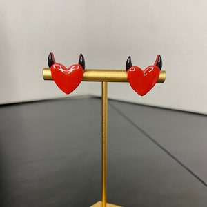 May include: A pair of red heart-shaped earrings with black devil horns on a gold stand.