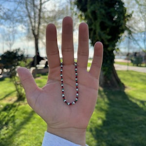 Puede incluir: Un collar de cuentas rojas, blancas y verdes. El collar se muestra en una mano abierta, sobre un fondo de hierba verde y árboles. Las cuentas son pequeñas y están espaciadas uniformemente.