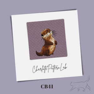 Peut inclure: Motif de point de croix représentant une loutre brune debout sur un fond violet. Le texte "Charlotte Pattern Lab" est écrit en cursive en dessous, avec "CB41" en dessous. L'image est sur fond blanc.