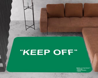 May include: A rectangular green rug with rounded corners, featuring the words "KEEP OFF" in large white capital letters. The rug is placed on a concrete floor, with a brown sofa in the background.