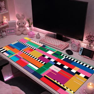 May include: A colorful rectangular desk mat with a geometric design in shades of blue, orange, green, purple, and yellow. The mat is placed on a white desk, with a computer monitor, keyboard, and other accessories visible in the background.