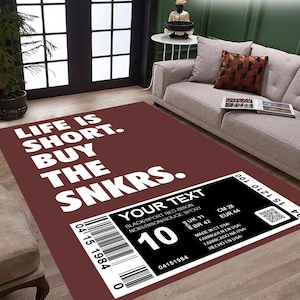 Pode incluir: Um tapete marrom com o texto "LIFE IS SHORT. BUY THE SNKRS." em branco. O tapete também possui uma etiqueta preta e branca com o texto "YOUR TEXT BLACK/SPORT RED-BISON NOIR/BISON/ROUGE SPORT 10 UK 11 CM 28 BR 42 EUR 44 04151984 MADE IN CT USA FABRIQUEN EN USA FABRICADO NA USA HECHO EN USA!"