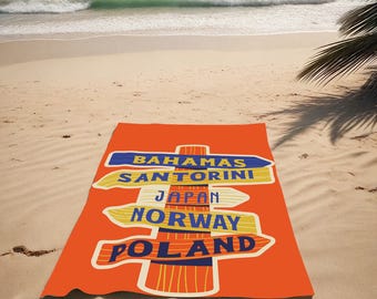May include: An orange beach towel with a signpost design featuring the names of travel destinations: Bahamas, Santorini, Japan, Norway, and Poland. The towel is on a sandy beach with the sea in the background.