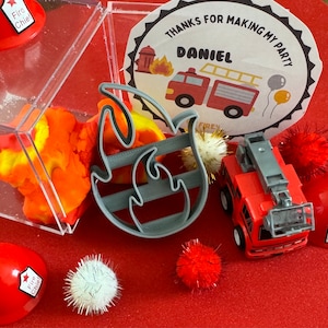 Peut inclure: Un camion de pompiers rouge jouet avec une échelle, un emporte-pièce en forme de flamme de feu gris, un récipient de pâte à modeler orange et jaune, et un chapeau de chef des pompiers rouge avec une étoile et les mots "Fire Chief" dessus. Il y a aussi des pompons rouges et blancs et une étiquette de remerciement blanche avec les mots "Merci d'avoir fait de mon anniversaire Daniel" dessus.