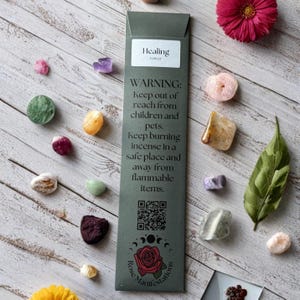 May include: An assortment of colorful crystals and a green incense package with the text "Healing Vetiver" and a warning label. The crystals are various shapes and sizes, scattered on a light wood-grain surface. A pink flower and a green leaf are also present.