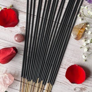 May include: A collection of black incense sticks with gold-colored bases, arranged on a light wood-grain surface. Red rose petals, small white flowers, and crystals surround the incense. The overall aesthetic is natural and serene.