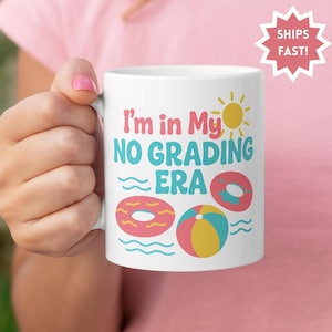 Könnte beinhalten: Weiße Keramik-Tasse mit dem Text "I'm in My NO GRADING ERA" in Türkis und Rosa. Die Tasse zeigt Illustrationen einer Sonne, zwei Donuts und einen Strandball. Die Tasse wird von einer Person mit rosa Nagellack gehalten.