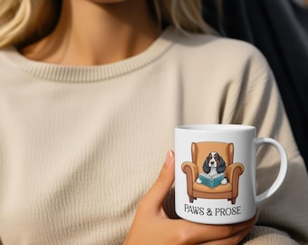 May include: White ceramic mug featuring a cartoon dog in a brown armchair reading a book. The mug has the text "PAWS & PROSE" in black. The dog is black, white, and brown.