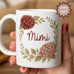 Op de afbeelding: Witte keramische mok met de naam "Mimi" in een sierlijk lettertype, omringd door een bloemenkrans van bordeauxrode en blushkleurige hortensia's en groene bladeren. De mok heeft een wit handvat en een "SHIPS FAST!" sticker.
