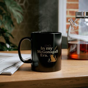 May include: Black ceramic mug with the text "In my McGonagall Era." in gold lettering. The mug features a cat illustration, glasses, and a feather. Steam rises from the mug, suggesting a hot beverage.