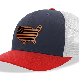 May include: A navy blue and red trucker hat with a white mesh back. The hat features a brown leather patch in the shape of the United States with a black and white American flag design.