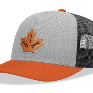 May include: A grey and orange baseball cap with a black mesh back. The cap has a leather patch with a maple leaf design and the text "6IX". The peak is orange, and the top button is orange.