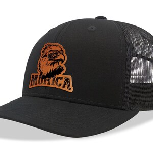 Op de afbeelding: Zwarte truckercap met een bruine leren patch met een adelaar die een zonnebril draagt en het woord "MURICA" in een gestileerd lettertype.