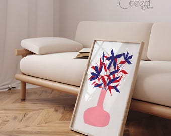 May include: A framed art print featuring a pink vase with red and blue floral elements. The artwork is displayed against a neutral-toned background, leaning against a beige sofa. The print has a light wooden frame.