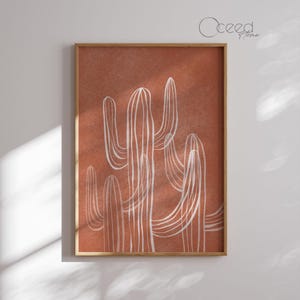 May include: A framed art print featuring a stylized illustration of cacti. The artwork showcases white line drawings of cacti against a warm, terracotta-colored background. The frame is a natural wood tone, adding to the earthy aesthetic. The print is suitable for home decor.