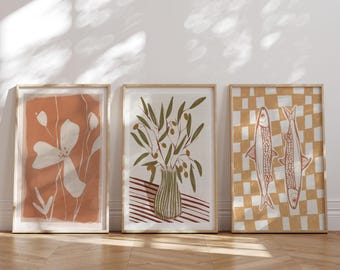 Könnte beinhalten: Drei gerahmte Kunstdrucke mit minimalistischer Ästhetik. Die Drucke zeigen botanische und Fischillustrationen in gedämpften Tönen von Orange, Weiß und Grün. Die Rahmen sind in Naturholz gehalten und unterstreichen den erdigen Charakter der Kunstwerke.
