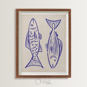 May include: A framed print featuring two stylized blue fish illustrations on a beige background. The fish are depicted in a simple, linocut style, with one upright and the other inverted. The frame is a warm brown color.