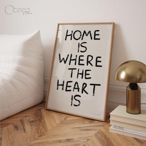May include: A framed print with the text "Home is where the heart is" in black lettering on a white background. The print is sitting on a wooden floor with a gold lamp and a stack of books.