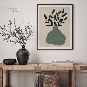 K&ouml;nnte beinhalten: Eine Schwarzwei&szlig;illustration einer Vase mit Bl&auml;ttern in gr&uuml;ner Farbe. Die Illustration ist gerahmt und h&auml;ngt an einer wei&szlig;en Wand. Im Vordergrund steht ein Holztisch mit einer gewebten Tischplatte.