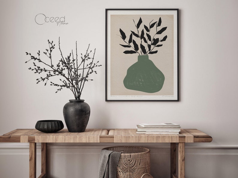 K&ouml;nnte beinhalten: Eine Schwarzwei&szlig;illustration einer Vase mit Bl&auml;ttern in gr&uuml;ner Farbe. Die Illustration ist gerahmt und h&auml;ngt an einer wei&szlig;en Wand. Im Vordergrund steht ein Holztisch mit einer gewebten Tischplatte.