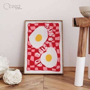 May include: A framed art print featuring two sunny-side-up eggs on a red and pink checkered background. The words "SUNNY SIDE UP" are printed in white. The print is in a wooden frame and is displayed on a wooden floor.