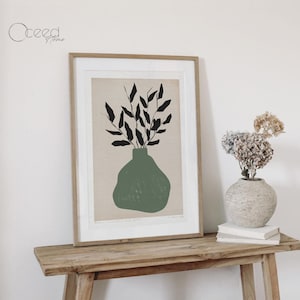 K&ouml;nnte beinhalten: Ein gerahmter Druck einer minimalistischen Illustration einer gr&uuml;nen Vase mit schwarzen Bl&auml;ttern. Der Druck h&auml;ngt an einer wei&szlig;en Wand &uuml;ber einem Holztisch.