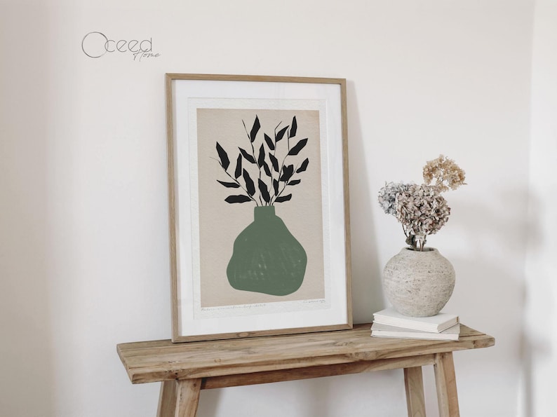 K&ouml;nnte beinhalten: Ein gerahmter Druck einer minimalistischen Illustration einer gr&uuml;nen Vase mit schwarzen Bl&auml;ttern. Der Druck h&auml;ngt an einer wei&szlig;en Wand &uuml;ber einem Holztisch.