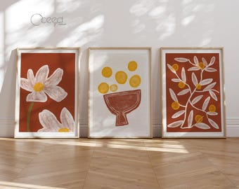 Könnte beinhalten: Drei gerahmte Kunstdrucke im minimalistischen Stil. Der erste Druck zeigt eine weiße Blume auf terracottafarbenem Hintergrund. Der zweite Druck zeigt eine weiße Schale gefüllt mit gelben Kreisen auf weißem Hintergrund. Der dritte Druck zeigt einen weißen Zweig mit gelben Kreisen auf terracottafarbenem Hintergrund.