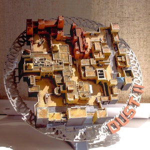 May include: A detailed model of a city layout, with buildings and structures in shades of brown, tan, and grey. The model is set within a circular, openwork frame. The word "DUST II" is displayed in orange letters.