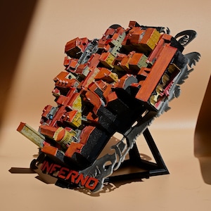 May include: A three-dimensional model of a dense city layout with orange-red rooftops and yellow building facades, mounted on a black stand. The base features the word INFERNO in bold red lettering, with decorative grey metal accents on the sides.