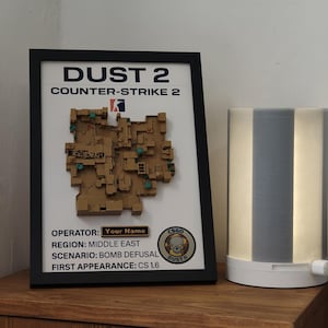 May include: Framed artwork featuring the Dust 2 map from Counter-Strike 2. The map is a 3D relief in shades of brown and green, set against a white background. The frame is black, and the text includes game details.