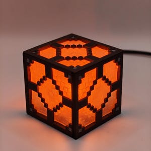 Op de afbeelding: Een kubusvormige lamp met een pixelachtig ontwerp, dat doet denken aan de esthetiek van een videogame. De lamp heeft een donkerbruin frame met een oranje gloed van binnenuit. Het ontwerp heeft geometrische patronen, waardoor een warm, omgevingslicht ontstaat.