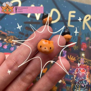 May include: A small, orange cat-shaped figurine with black eyes and stripes on its head. The figurine is held in a hand, with a background of a colorful illustration and the word "DER". The image also includes the text "ko-fi.com/memoruri" and "memoruri".