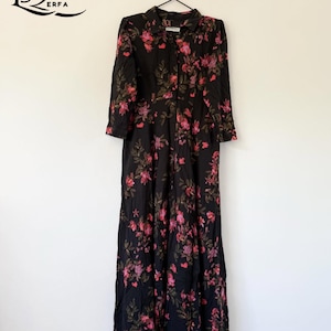 May include: A long black dress featuring a floral design of pink flowers and green leaves. The dress has long sleeves, a collared neckline, and buttons down the front. It is suspended from a metal hanger.